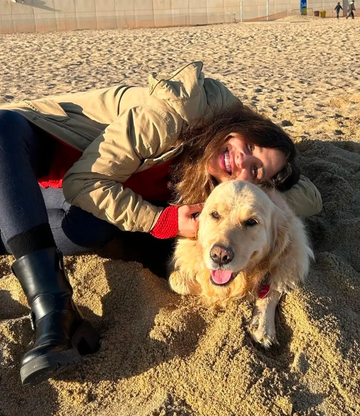 Mariana tumbada en la arena abrazando a un golden retriever bajo el sol de invierno en Barcelona